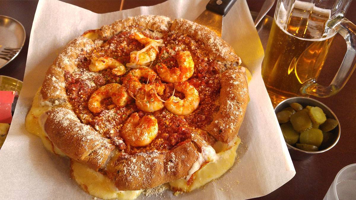 pizza topped with shirmps with cheese oozing in the bottom with a side of pickles paired with a pint of beer in Chicago, USA